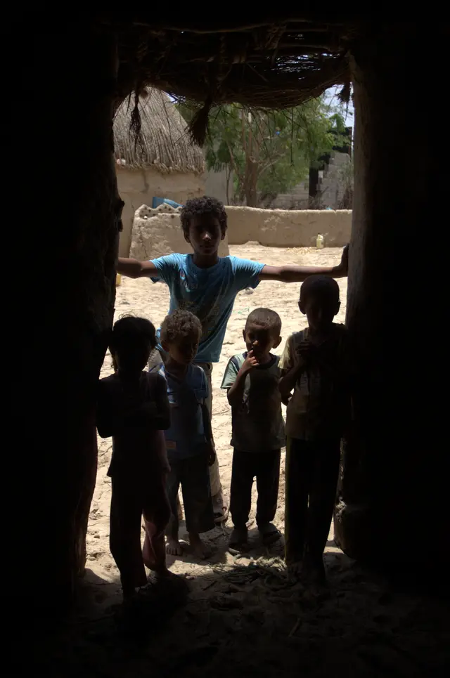 Silhouettes of children standing in a dark doorway, with sunlit houses beyond.