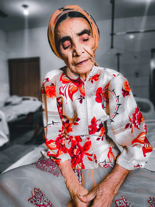 An elderly woman in a red floral blouse and orange headscarf sits with her eyes closed and hands folded in her lap.