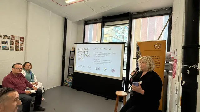 A woman speaks into a microphone beside a screen showing the Journeys of Courage programme of the Human Rights Festival 2026.
