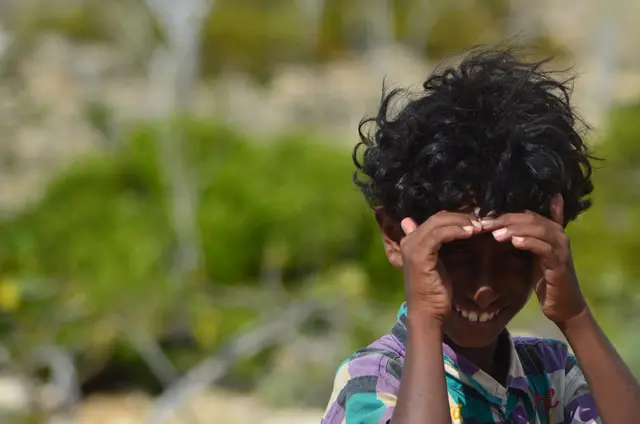 A laughing boy with tousled hair covers his eyes with his hands.