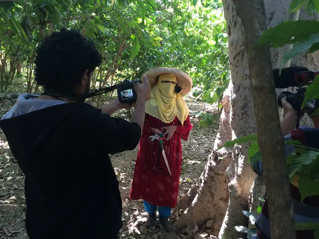 A man films with a video camera as a person in a wide straw hat and face covering stands among trees.
