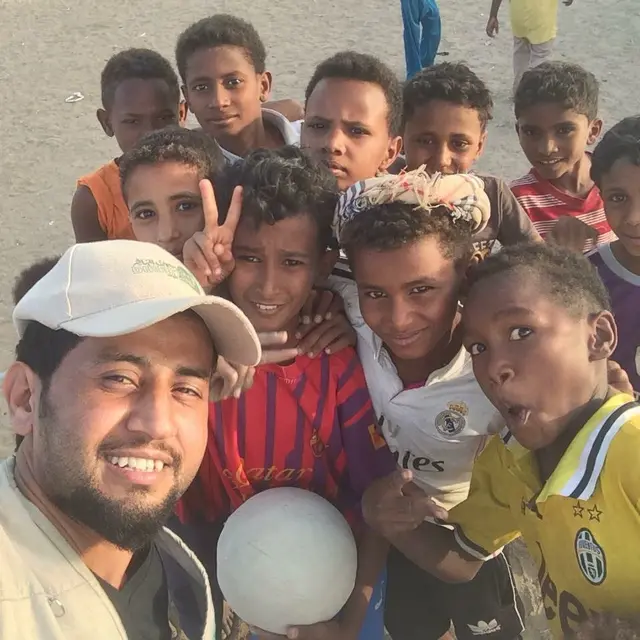 A smiling man in a white cap takes a selfie with a group of boys, one holding a football.
