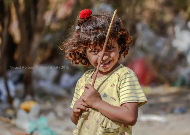 A smiling girl with curly hair and a red hair clip holds a thin stick outdoors.
