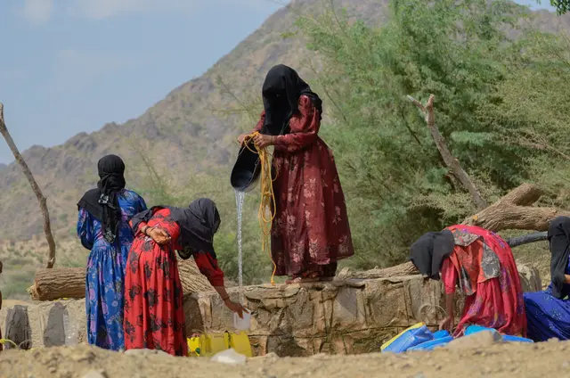 Women in long dresses and headscarves draw water at a stone well, with a mountain behind them.